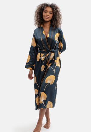 Woman with curly hair wearing a dark robe with large yellow floral patterns, standing barefoot against a plain background.