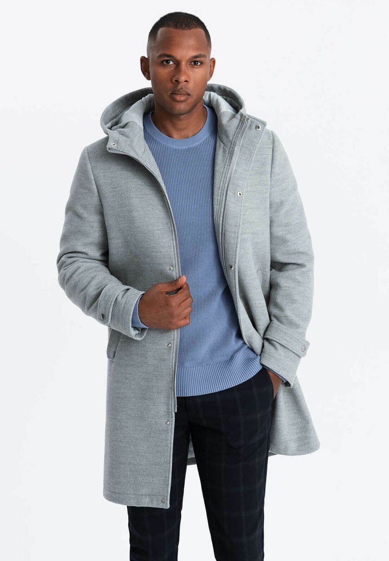 Abrigo gris con capucha, con cierre frontal con cremallera y cierre a presión. Lleva encima un suéter de canalé azul y pantalones de cuadros oscuros.