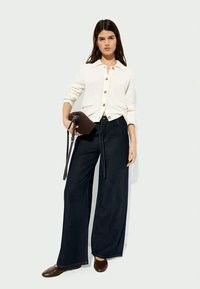Woman wearing a white buttoned cardigan, wide-leg dark jeans, brown shoes, and holding a small brown handbag against a plain background.