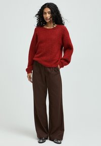 Rotem, strukturiertem Pullover mit rundem Ausschnitt, kombiniert mit hellbraunen Hosen mit schmalen Streifen. Das Model steht vor einem hellgrauen Hintergrund.