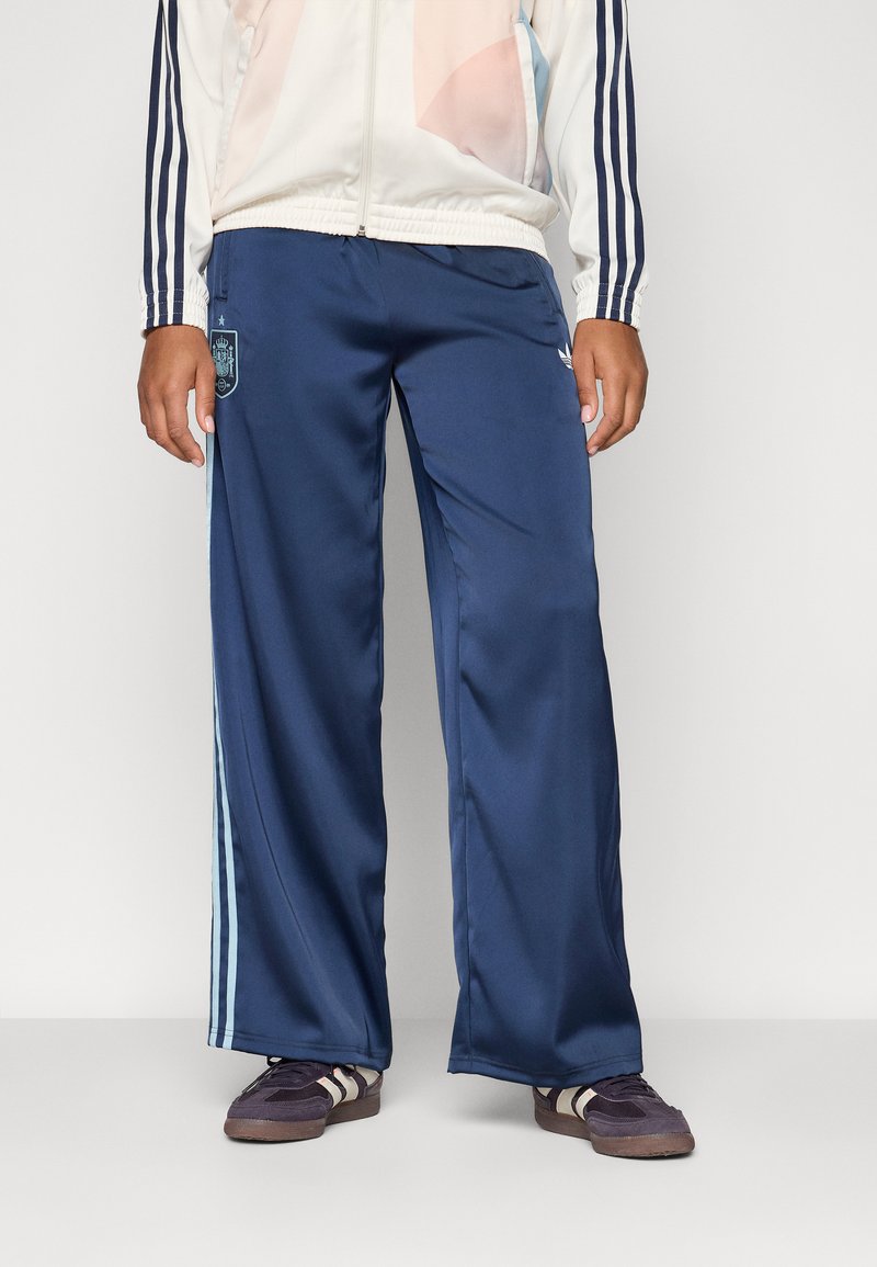 Jūrų mėlynos satino sportinės kelnės su šviesiai mėlynomis šoninėmis juostelėmis, kairiajame šlaunyje turi logotipą ir yra laisvos formos. Derinamos su tamsiais sportbačiais.