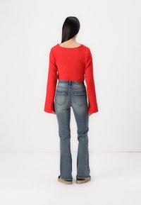 Rød kort bluse med lange ermer, paret med tettsittende blå bootcut jeans. Stoffet på blusen er teksturert, og jeansene har baklommer.