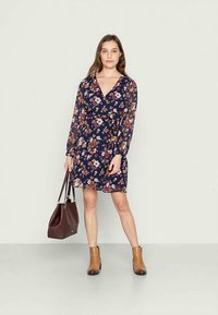 Vestido floral azul marinho com um design envolvente, mangas longas transparentes e decote em V. Usado com botas de tornozelo castanhas e segurando uma bolsa burgundy.
