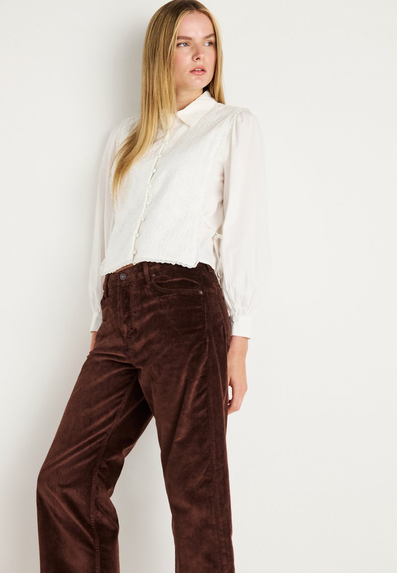 Witte blouse met hoge hals en knoopdetails, voorzien van kanten accenten en lange mouwen, gecombineerd met bruine corduroy wijde broek.