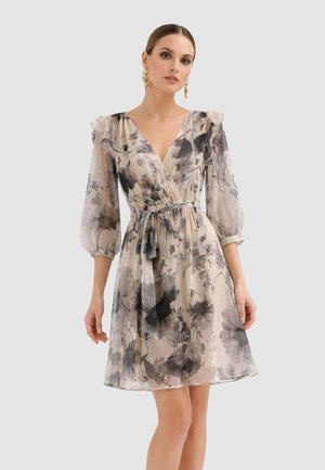 Donna che indossa un abito floreale trasparente al ginocchio con maniche a sbuffo a tre quarti, scollo a V e cintura in vita, su sfondo chiaro uniforme.