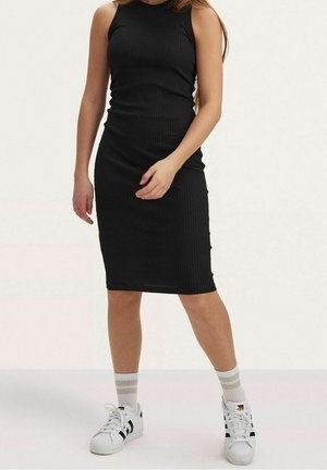 Femme portant une robe noire côtelée sans manches, longueur genou, assortie à des baskets blanches Adidas et des chaussettes blanches à rayures beiges.