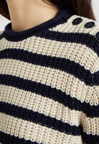 Pull en tricot présentant des rayures alternées bleu marine et crème, tissu texturé, et encolure ronde avec des boutons décoratifs noirs sur l'épaule.