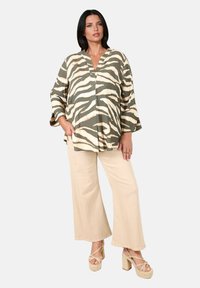 Femme aux cheveux bruns portant un haut zèbre rayé vert et beige, un pantalon large beige et des talons compensés beiges, se tenant avec une main dans sa poche.