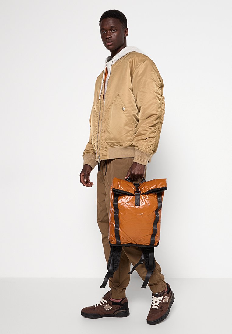 Zaino in nylon marrone con spallacci neri, chiusura a rullo e texture lucida. Indossato con una giacca bomber beige e pantaloni marroni.