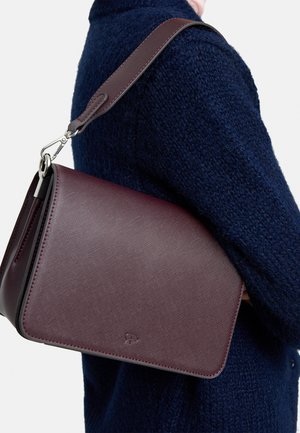 Kobieta w ciemnoniebieskim teksturowanym płaszczu niesie na ramieniu usztywnioną bordową skórzaną torbę na ramię z szerokim paskiem.