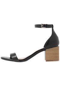 Sandale en cuir noir avec un talon carré en bois. Équipée d'une bride à la cheville et d'une bande frontale minimaliste. Texture lisse, design épuré.