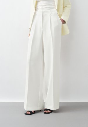 Persona con pantalones blancos de tiro alto y corte ancho, blazer beige, top blanco y tacones abiertos negros, posando frente a un fondo blanco liso.