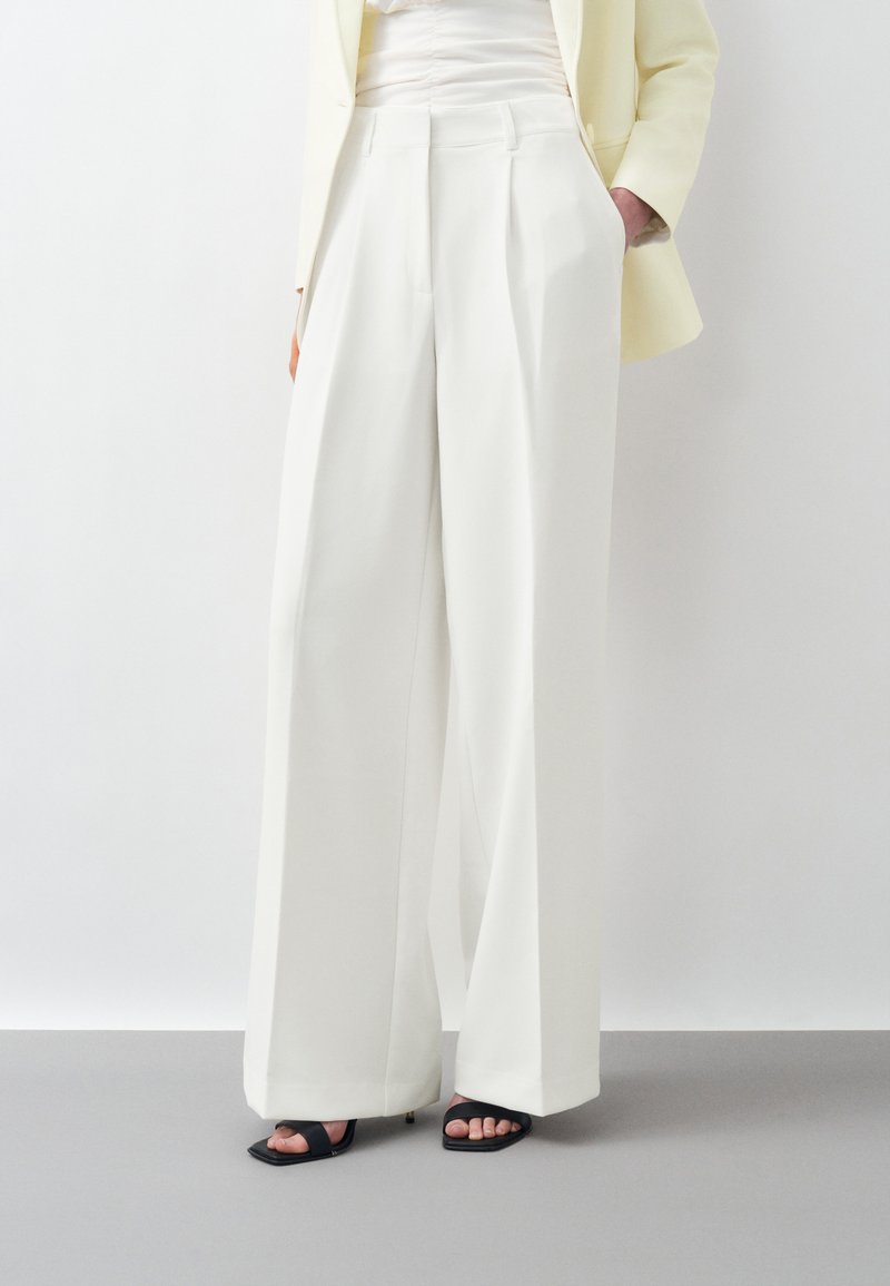 Personne portant un pantalon blanc taille haute à jambes larges, un blazer beige, un top blanc et des talons noirs ouverts à l'avant, debout devant un fond blanc uni.