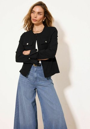 Femme aux bras croisés portant une veste noire boutonnée, des boucles d’oreilles créoles dorées et un jean large bleu clair sur fond blanc.