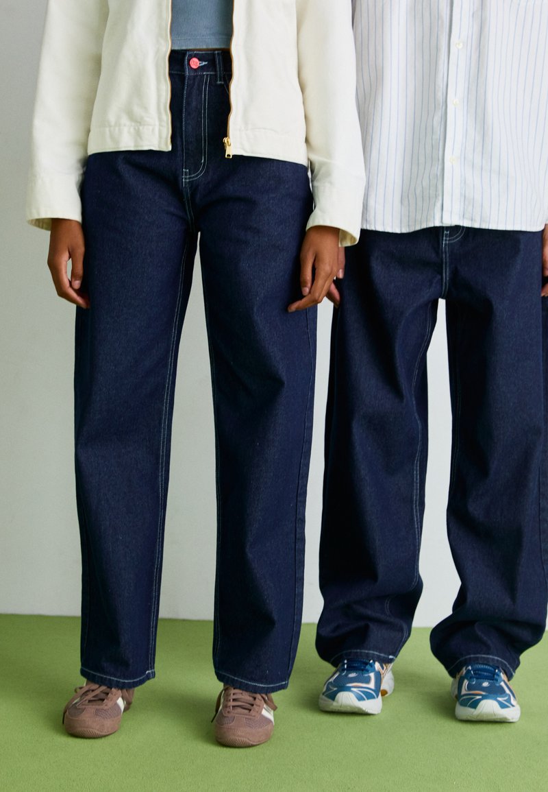Mørkeblå jeans med rette ben, med hvit søm, brukt sammen med beige og stripete skjorter. Brune og blå sneakers på føttene.