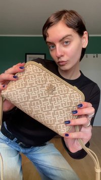 Beige portemonnee met een gestructureerd patroon en een logo, ritssluiting en een afneembare band, getoond in een handpositie.