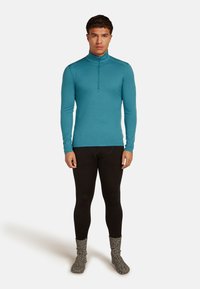 Man wearing a teal long-sleeve half-zip shirt, black base layer leggings, and speckled wool socks, standing against a plain background.