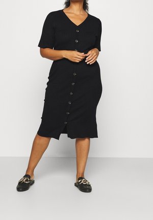 Femme portant une robe midi noire à col en V boutonnée avec des manches courtes, accompagnée de mocassins noirs à enfiler ornés d'un détail en chaîne dorée.