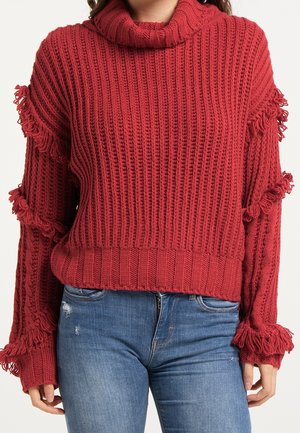 Femme portant un pull rouge en maille avec des détails à franges sur les manches et un jean bleu déchiré devant un fond clair uni.