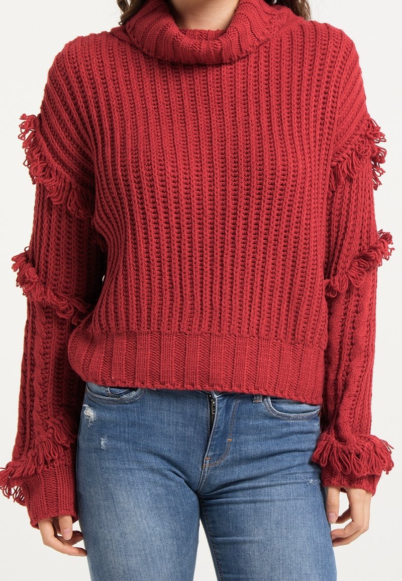 Femme portant un pull rouge en maille avec des détails à franges sur les manches et un jean bleu déchiré devant un fond clair uni.