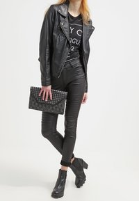 Chaqueta de moto de cuero negro sobre una camiseta gráfica negra, combinada con jeans ajustados negros y botas de tobillo. Sosteniendo un bolso de mano con studs.