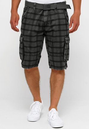 Mand iført sorte ternede cargoshorts med bælte og hvide sneakers, stående mod en ensfarvet lys baggrund.