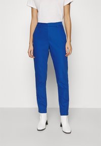 Kvinna som bär ljusblå smalpassande byxor, vit kortärmad t-shirt och vita ankelskorsor, stående framför en enkel vit bakgrund.