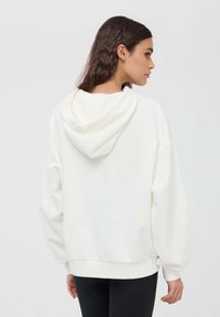 Felpa bianca con una consistenza morbida, dotata di cappuccio con coulisse, maniche ampie e polsini a costine. Si abbina a pantaloni scuri.