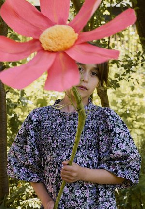 Noor inimene lillelise kleidiga, kes hoiab pikka varrega suurt roosat lille, seisab õues roheliste puude ja päikesevalguse keskel.