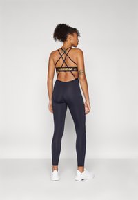 Meriininen kompressiobodysuit ristiinlasketuilla takaolkanauhoilla, jossa on kultainen NEBBIA-logo. Silkkinen pinta ja istuva malli urheilukäyttöön.