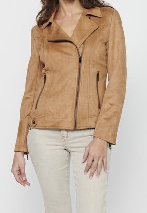 Mujer con chaqueta de ante marrón claro con cremallera frontal y bolsillos con cremallera, combinada con pantalones beige claro.
