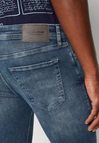 Jeans de denim en azul oscuro con un ligero desgaste, que presentan un parche de cuero etiquetado "Jack & Jones" y un diseño estándar de bolsillo trasero.