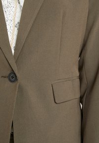 Veste olive en tissu structuré, dotée d'un revers cranté, d'une fermeture à un bouton et d'une poche latérale. Fini texturé.