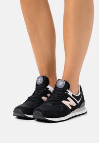 Ben som bär svarta och vita New Balance 574 sneakers med en beige "N"-logotyp, stående på en enkel ljus bakgrund.