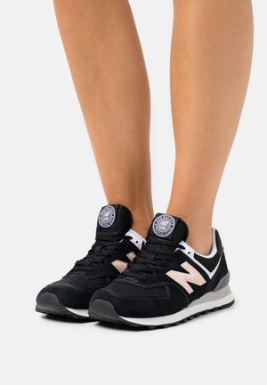 Ben som bär svarta och vita New Balance 574 sneakers med en beige "N"-logotyp, stående på en enkel ljus bakgrund.