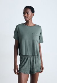 Kvinna klädd i en lös kortärmad grön topp med ett bröstficka och gröna shorts med prickar och volanger som står mot en enfärgad bakgrund.