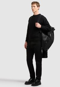 Jeune homme vêtu entièrement de noir, tenant une veste noire sur l'épaule sur un fond gris uni.