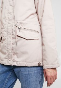 Ljusrosa jacka med dragkedjefickor och snoddar, bärs med blå denimjeans, visar sidan och nedre delen av kroppen på en person.