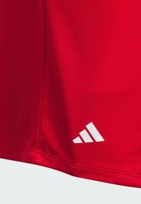 Czerwona sportowa koszulka z gładkiego materiału, z białym logo Adidas na dole i przeszyciami wzdłuż szwów.