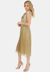 Metaliczna złota sukienka midi z teksturowanej tkaniny, z falbankami na ramionach i swobodnym krojem, zestawiona z cielistymi sandałami na wysokim obcasie.