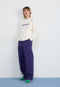 Krema pulover s kapuco z mornarsko modrim logotipom, v kombinaciji z ohlapnimi vijoličnimi hlačami in zelenimi supergami z vijoličnimi črtami na modri podlagi.