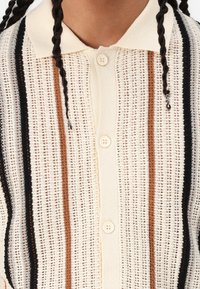 Strickjacke mit cremefarbener Basis, die vertikale Streifen in Schwarz, Braun und Dunkelblau aufweist. Verfügt über eine Knopfleiste vorne und einen strukturierten Stoff.