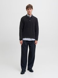 Giovane uomo con maglione nero a maglia sopra camicia a righe, pantaloni neri a gamba larga e scarpe eleganti nere, in piedi davanti a uno sfondo bianco.
