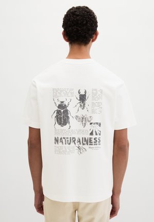 Fehér pólót viselő férfi, fekete rovar- és méhvázlatokkal, valamint a természetességről szóló szöveggel a háton, fehér háttér előtt áll.