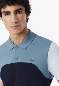 Paita polo dengan desain blok warna biru dan navy, bahan bertekstur, leher dua kancing, dan logo buaya kecil yang disulam.