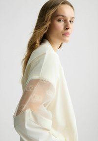 Weiße Bluse mit transparenten bestickten Ärmeln, die ein sich wiederholendes Logo-Muster aufweisen. Weiches Material und strukturiertes Kragen-Design mit umgeschlagenen Manschetten.