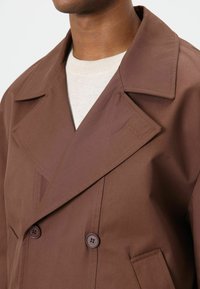Bruine double-breasted jas met grote revers, gestructureerde stof, twee bruine knopen en een zijzak. Gedragen over een lichtkleurige trui.