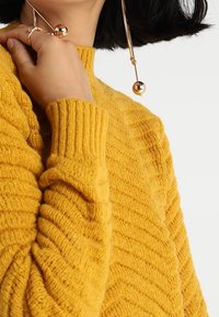 Žlutý pletený svetr s žebrovanou strukturou a vysokým límcem, s dlouhými rukávy. Doplňky zahrnují dlouhé zlaté náušnice s kulatými akcenty.