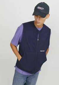 Veste matelassée marine avec une fermeture éclair, deux poches et le logo "Originals Studio". Portée sur un t-shirt violet et associée à un jean en denim clair.