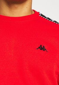 T-shirt en coton rouge avec un col rond côtelé, arborant un logo noir et des accents à motifs multicolores sur les épaules. Texture lisse.